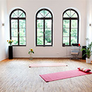 Foto: Großer hoher Raum mit weißen Wänden und 3 hohen Fenstern mit Rundbogen im Hintegrund. Im Vordergrund, mittig im Zimmer 2 Yogamatten auf Parkett.