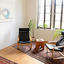 Foto: 2 Schwingstühle um einen kleinen Holzhockertisch auf Perser-Teppichläufer auf Parkettfußboden. Im Hintergrund 2 große Fenster linksseitig und rechtsseitig ein Wandbild.