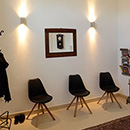 Foto: Foto: 3 Stühle neben Garderobenständer mit Jacke. Darunter ein Paar Schuhe. Mittig ein Bild an der Wand. Rechter Bildrand Durchgang zu den Praxen.