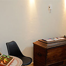 Foto: linker Bildrand Tisch mit Teebeutel-Sortiment. re. daneben Stuhl, re. danaben große Holztruhe. Darüber 2 Wandstrahler. Linker Bildrand oben eine alte Wanduhr.